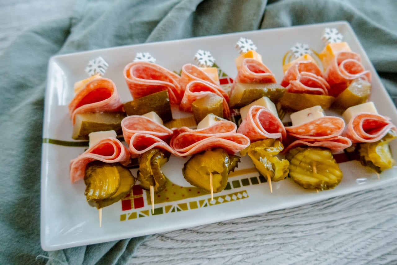A rectangular plate holds skewers with layers of folded salami, cubes of cheese, and sliced pickles, arranged neatly in rows. The plate rests on a green cloth.