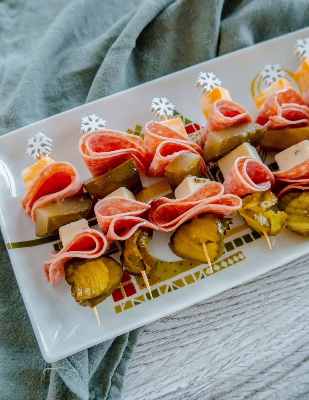 A rectangular plate displays skewers with slices of salami, cubes of cheese, and pickle slices, arranged in layers. The skewers are topped with decorative snowflake picks and placed on a patterned platter.