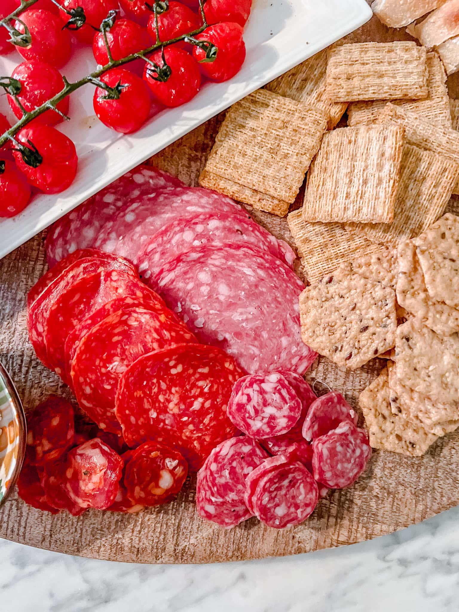 An overhead shot of a charcuterie board with vine tomatoes, Triscuit wheat crackers, hexagon seeded crackers, and assorted sliced cured meats including salami and pepperoni, arranged neatly on a wooden surface.