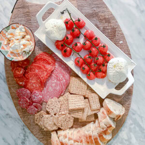 Overhead shot of a charcuterie board with sliced bread, assorted crackers, cured meats, cherry tomatoes on the vine, two balls of mozzarella, and a small bowl of soft cheese, arranged on a round wooden platter.