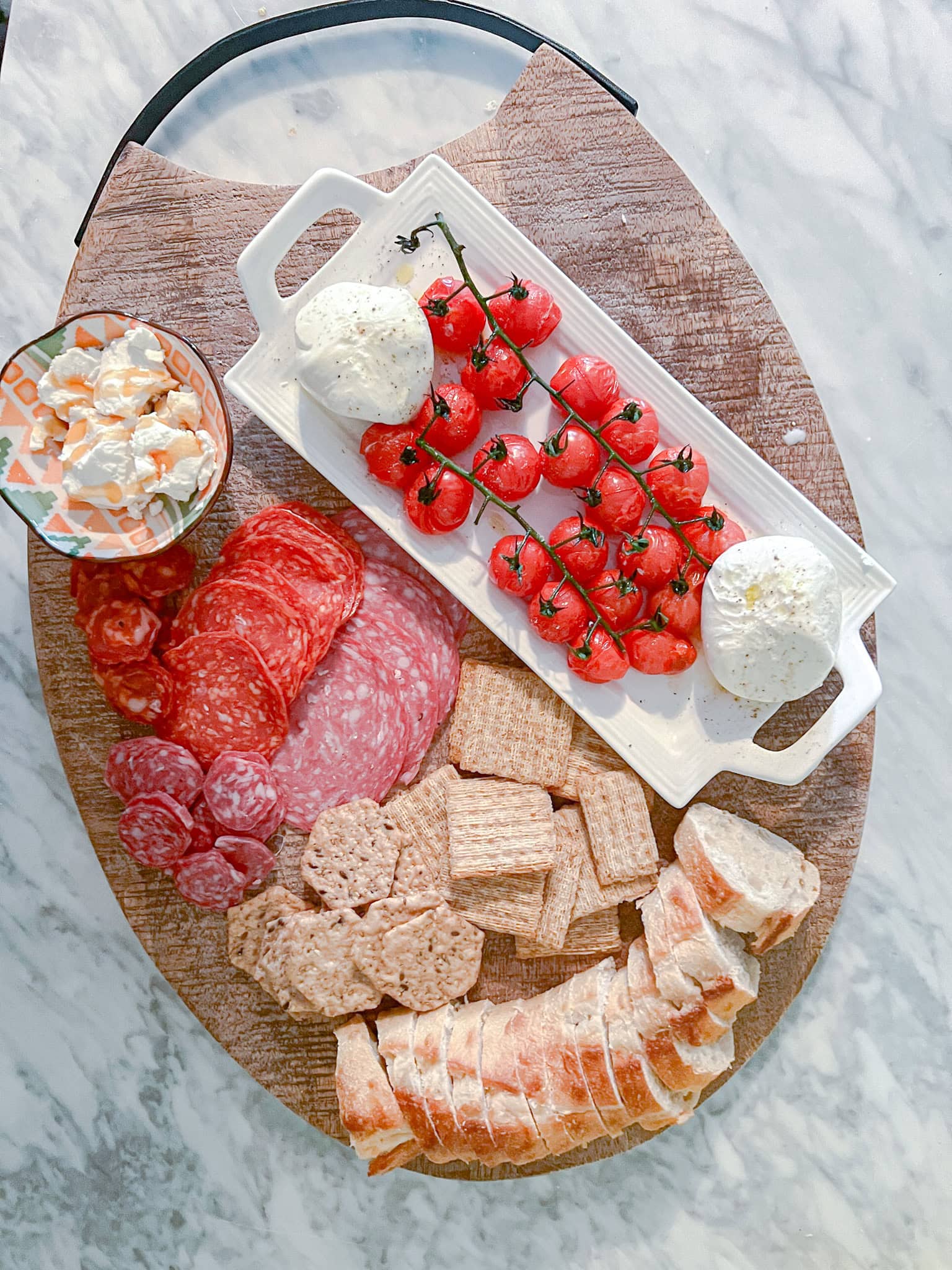 Overhead shot of a charcuterie board with sliced bread, assorted crackers, cured meats, cherry tomatoes on the vine, two balls of mozzarella, and a small bowl of soft cheese, arranged on a round wooden platter.
