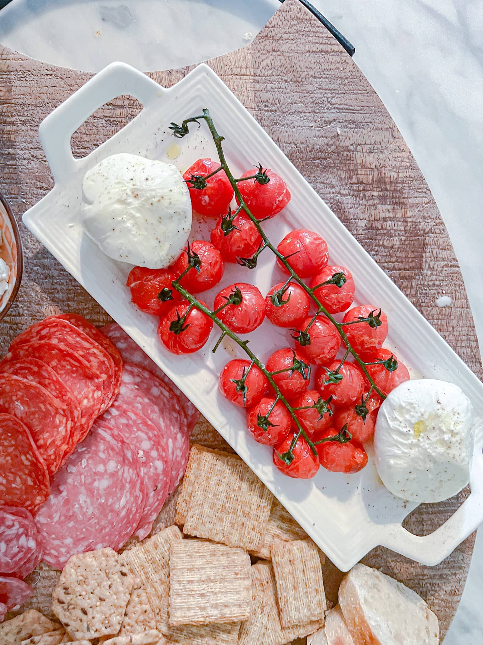Overhead shot of a platter with two balls of fresh mozzarella, a vine of cherry tomatoes, slices of salami, and assorted crackers arranged on a wooden board.