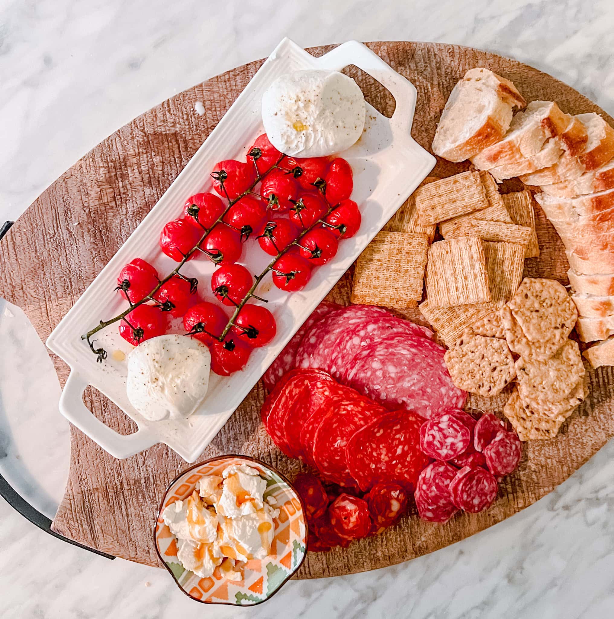 Overhead shot of a charcuterie board with sliced baguette, crackers, cured meats, a bunch of cherry tomatoes, mozzarella balls, and a small bowl of ricotta cheese, arranged on a wooden surface.