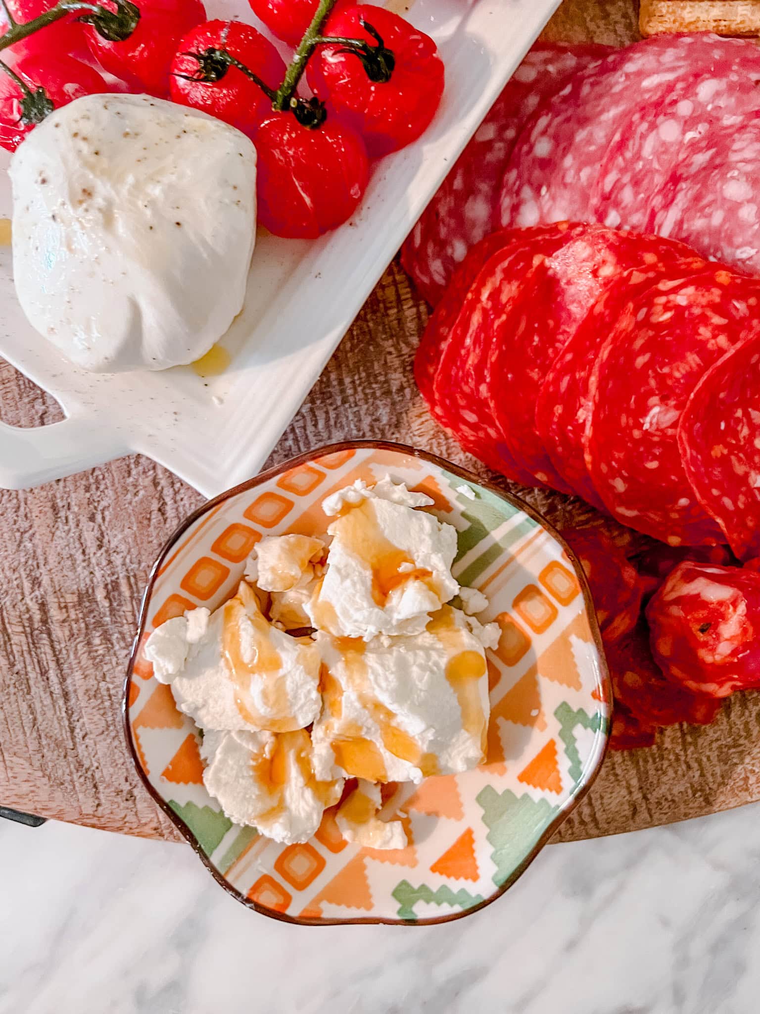 A close-up overhead shot of a small patterned bowl with ricotta cheese drizzled with honey sits next to sliced salami, mozzarella with olive oil, and roasted tomatoes on a wooden board.