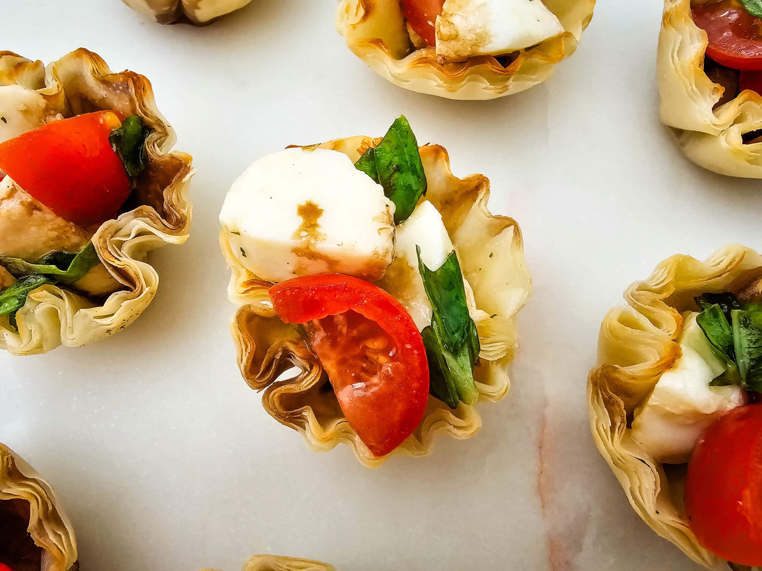 Overhead close-up of phyllo pastry cups filled with mozzarella, tomato slices, fresh basil, and a drizzle of balsamic glaze, arranged on a white surface.