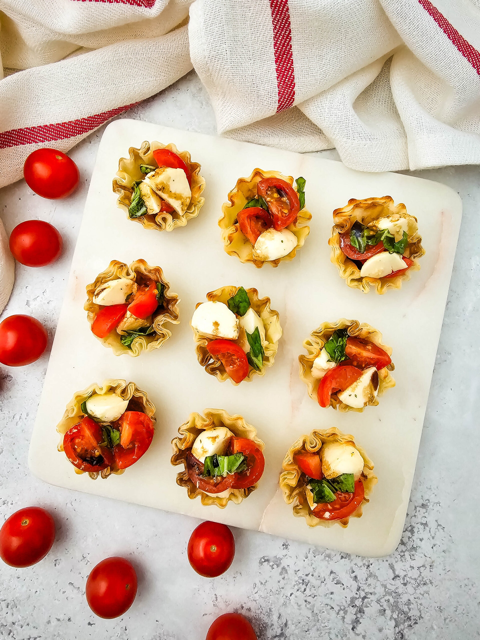 overhead shot of a marble board displaying nine bite-sized phyllo cups filled with cherry tomatoes, mozzarella, and basil. Whole cherry tomatoes and a white cloth with red stripes are scattered around the board.