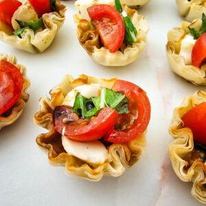 Mini phyllo pastry cups filled with sliced tomatoes, mozzarella cheese, fresh basil, and balsamic glaze, arranged on a white marble surface.