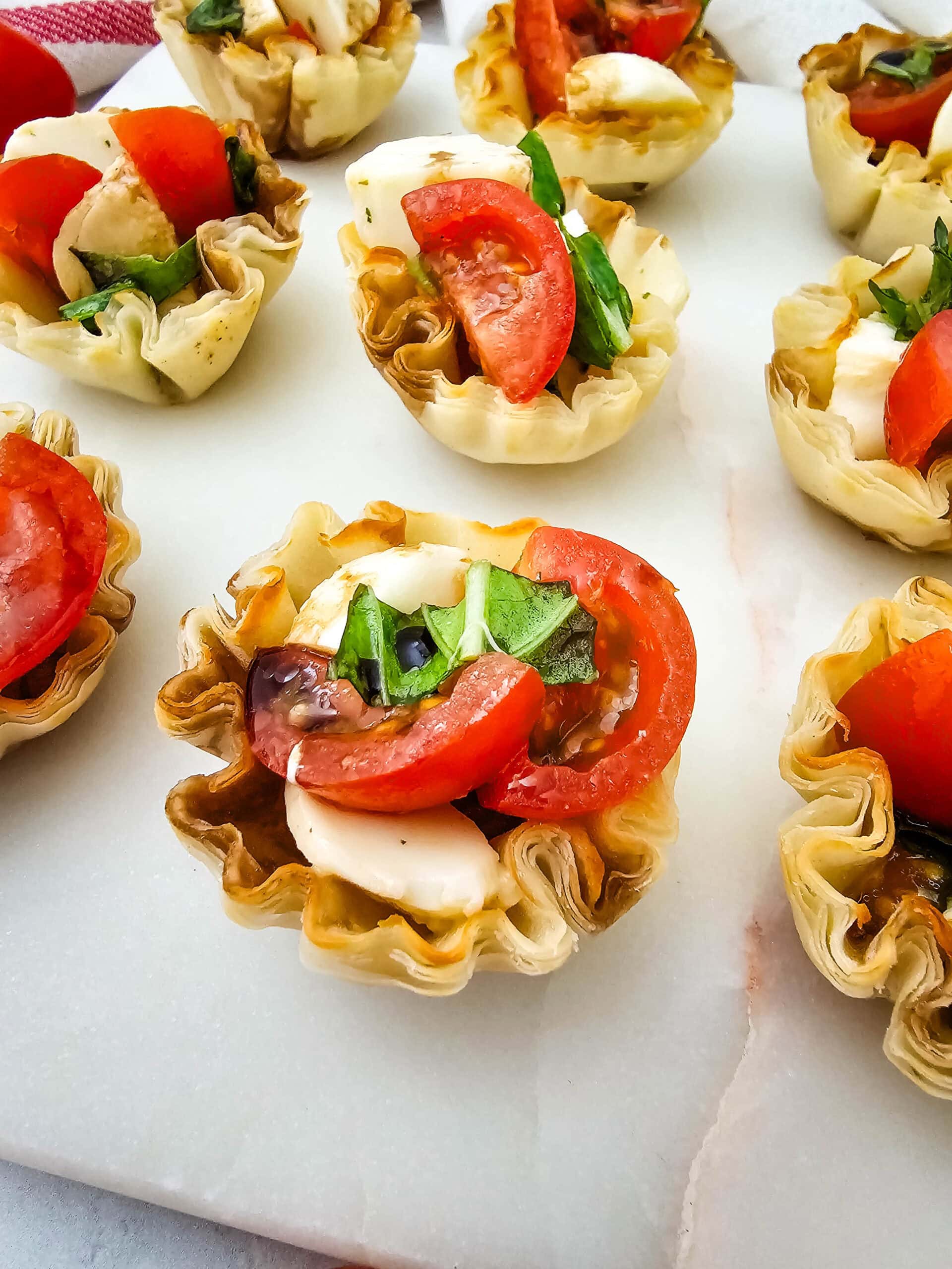 Mini phyllo pastry cups filled with sliced tomatoes, mozzarella cheese, fresh basil, and balsamic glaze, arranged on a white marble surface.