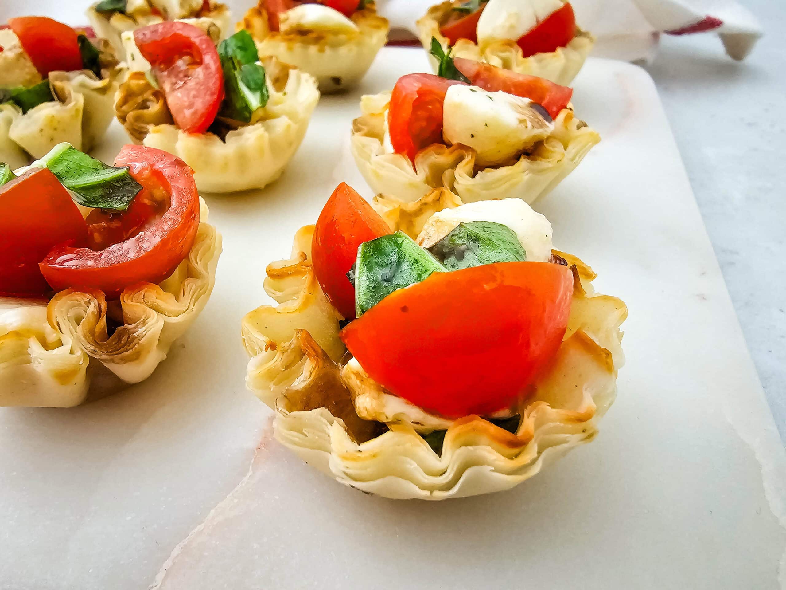 Close-up shot of mini phyllo pastry cups filled with chopped tomatoes, mozzarella balls, and fresh basil leaves, arranged on a white marble surface.