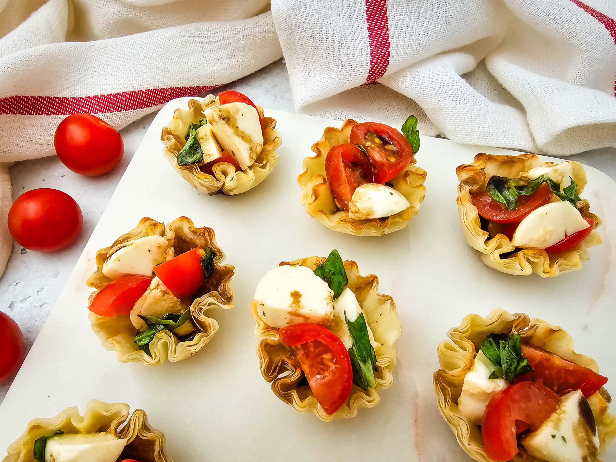 Mini phyllo cups filled with mozzarella, cherry tomatoes, fresh basil, and a drizzle of balsamic glaze, arranged on a white surface with a red-and-white striped cloth and whole cherry tomatoes in the background.