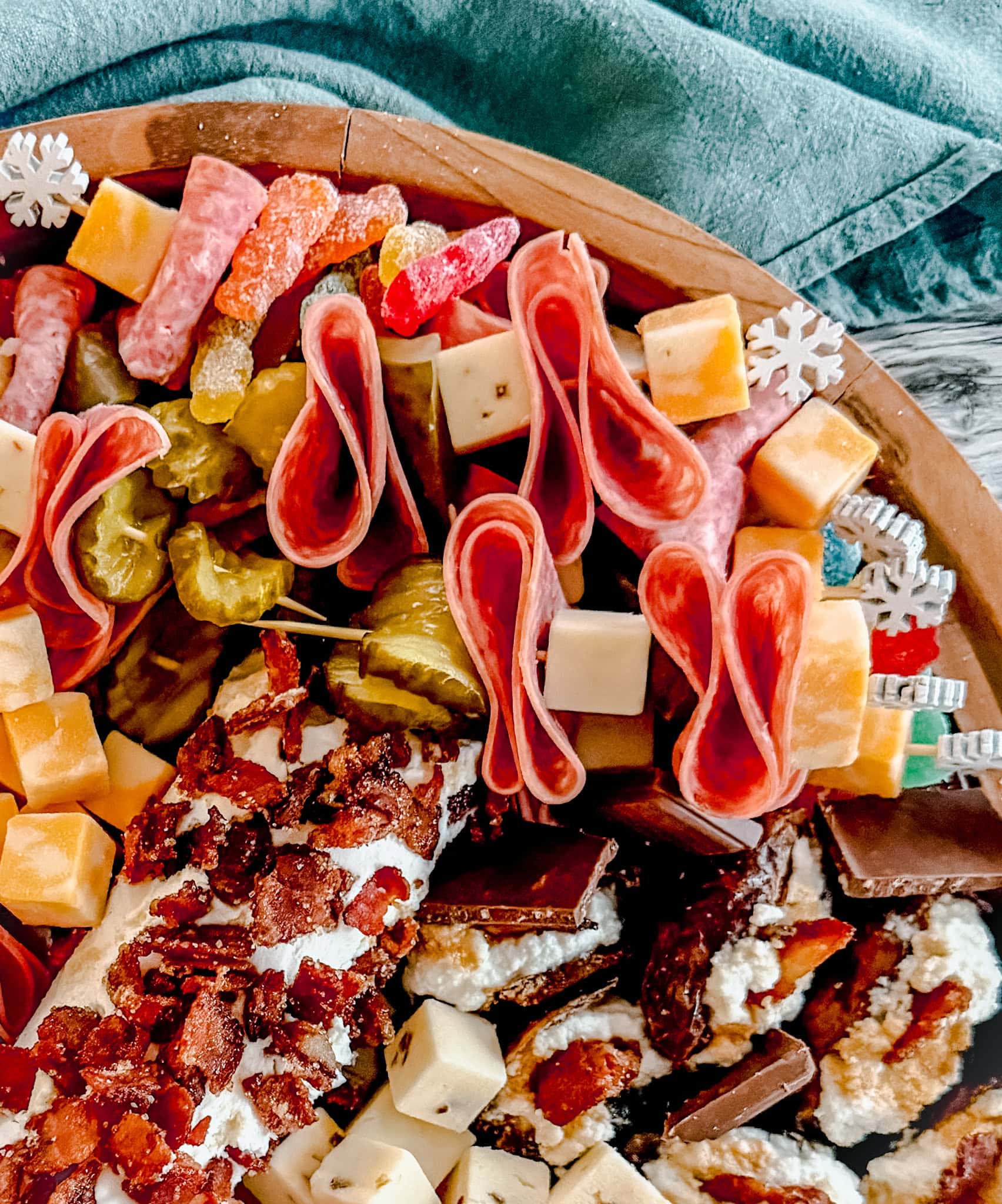 Overhead shot of a charcuterie board with rolled salami, cubed cheese, pickles, chocolate pieces, and a cheese log topped with bacon, set on a wooden tray with festive picks and a teal napkin nearby.