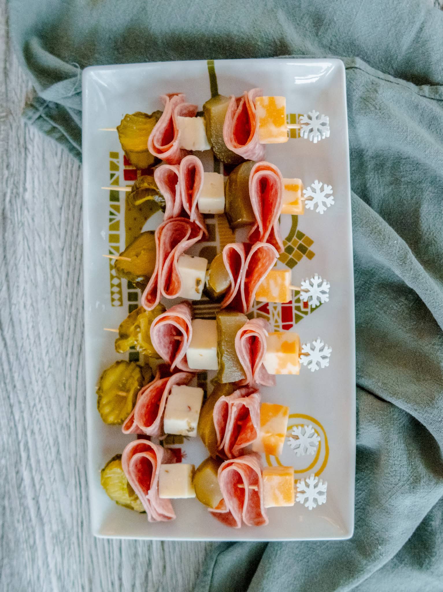 An overhead shot of a rectangular platter holds skewers with salami, cheese cubes, and pickle slices, arranged neatly in rows. The skewers have decorative snowflake picks, and the plate rests on a light gray fabric.