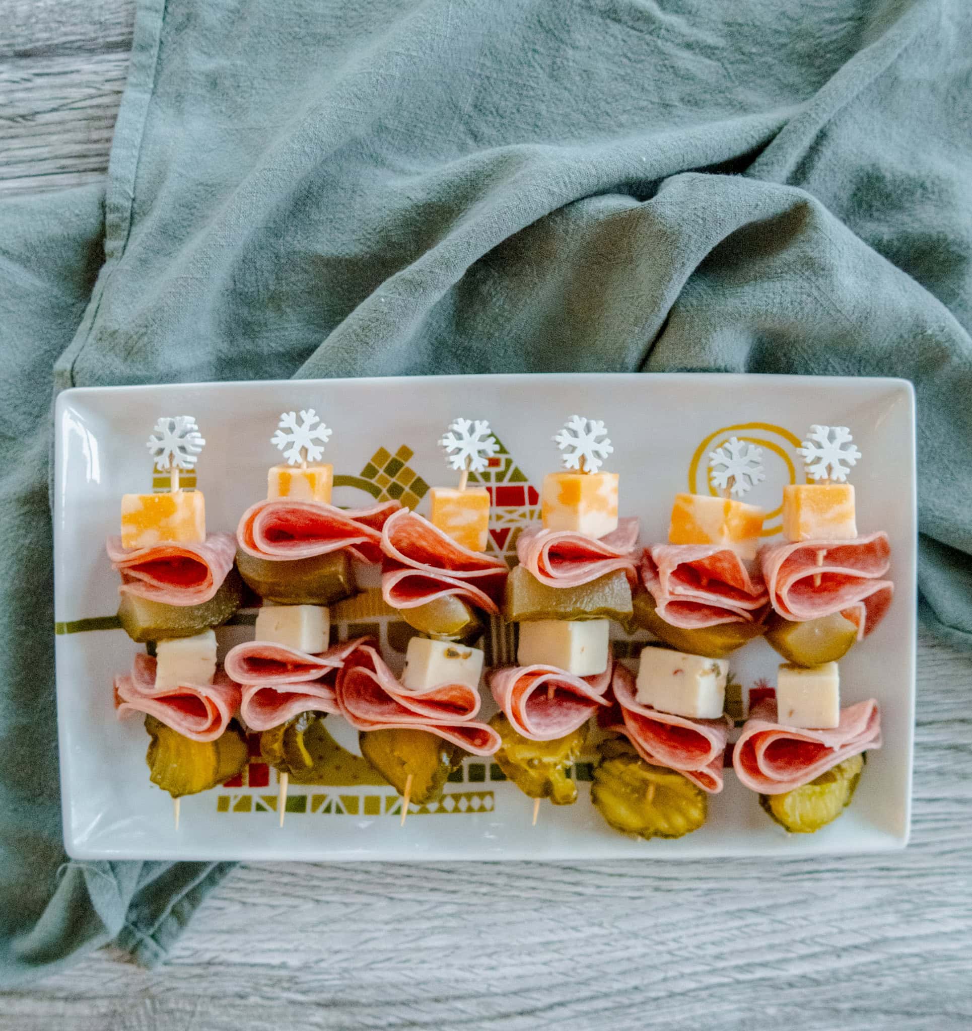 An overhead shot of a rectangular plate holds appetizer skewers with slices of pickles, cubes of cheese, and folded deli meat, each topped with a snowflake toothpick, on a wooden surface with a gray cloth nearby.