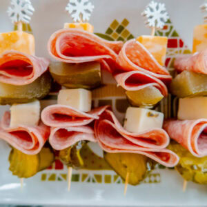 An overhead, close-up shot of skewers with slices of salami, cheese cubes, and pickle pieces arranged on a white plate decorated with a festive tree pattern; the skewers have white snowflake toppers.