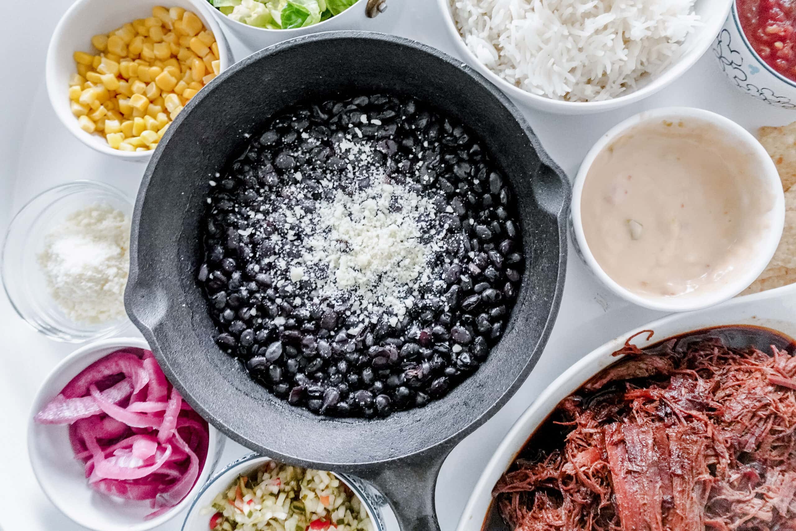 A cast iron skillet with black beans topped with cheese, surrounded by bowls of rice, corn, pickled onions, shredded meat, lettuce, sauce, salsa, and grated cheese—perfect for a Snug and Cozy Life gathering on a white surface.