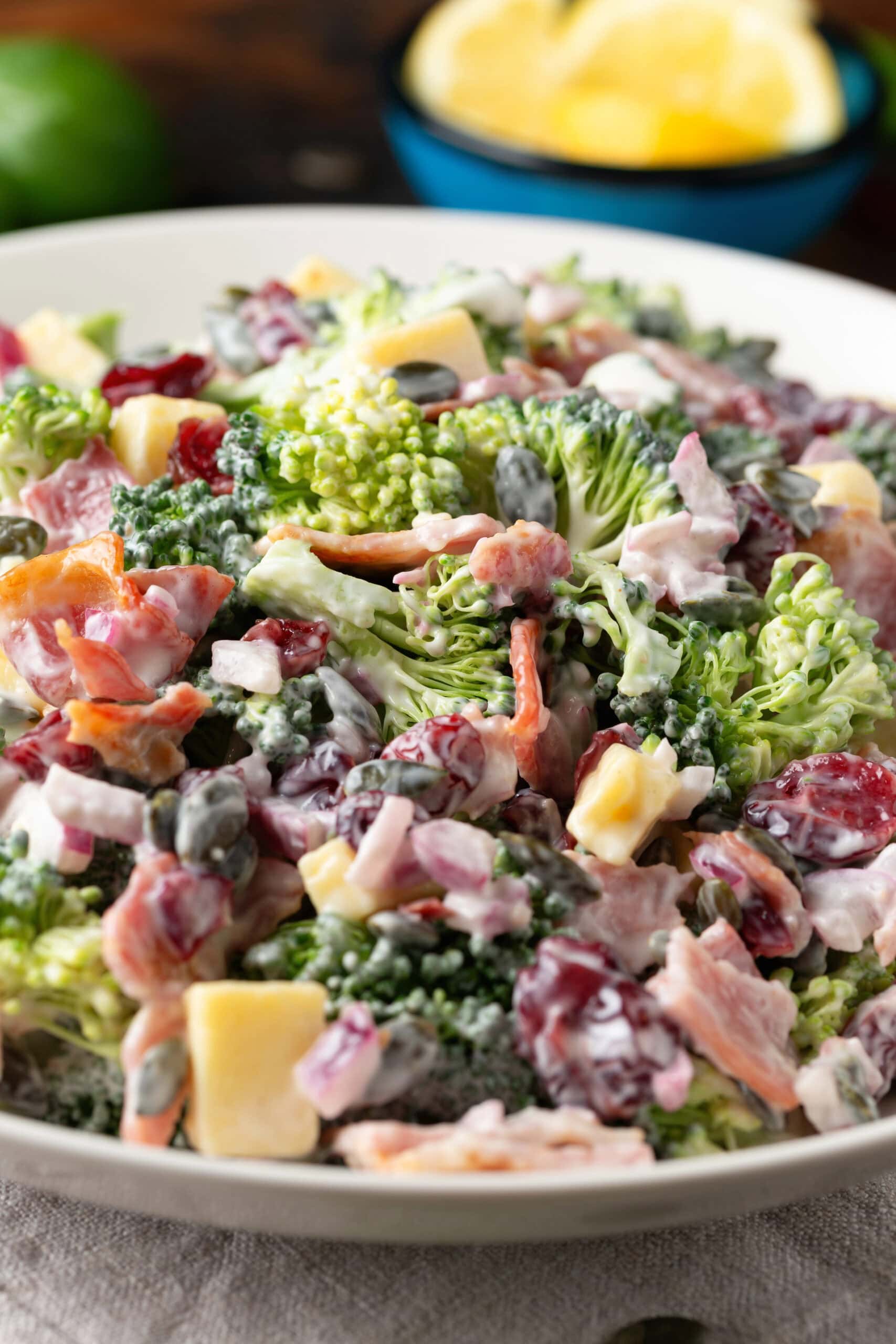 A close-up of a creamy broccoli salad&mdash;perfect for Easter side dishes&mdash;with chopped broccoli, cheese cubes, dried cranberries, sunflower seeds, and bacon pieces in a white bowl. A bowl of lemon wedges is blurred in the background.