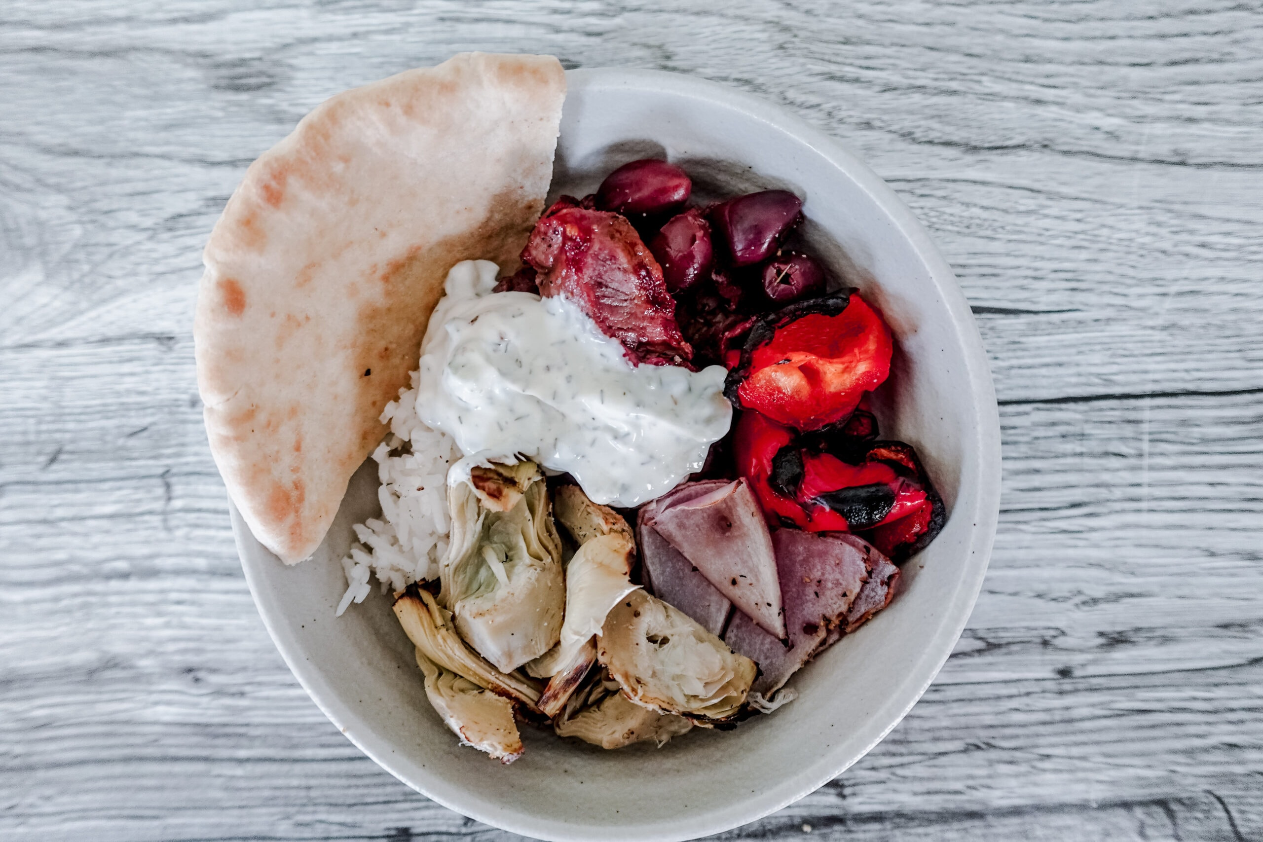 A bowl with pita bread, rice, grilled artichoke, roasted red peppers, black olives, sliced meat, and a dollop of tzatziki sauce, on a light wooden surface.