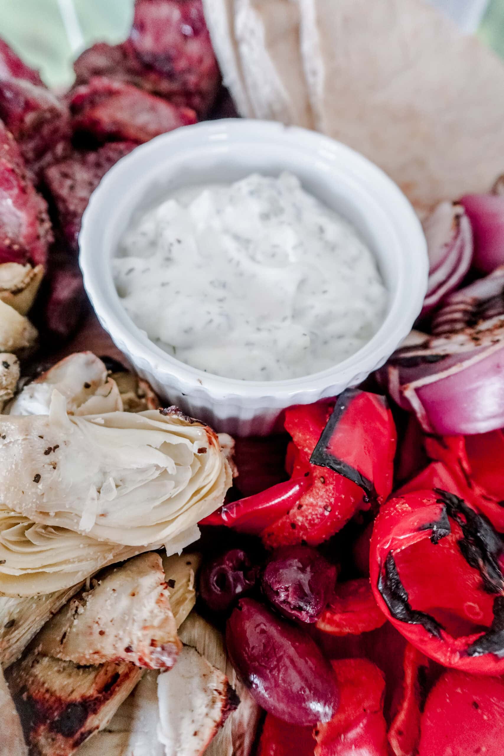A close-up of a Mediterranean platter featuring grilled red peppers, artichokes, olives, pita bread, red onions, and a small bowl of creamy tzatziki dip with herbs.