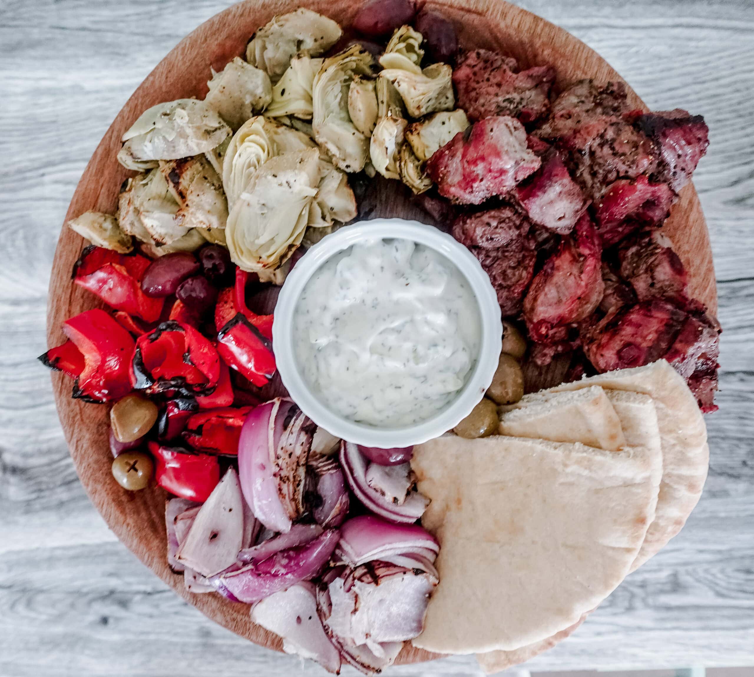 An overhead shot of a wooden platter with grilled steak, artichokes, red bell peppers, olives, red onions, pita bread, and a bowl of tzatziki sauce in the center, arranged in sections.
