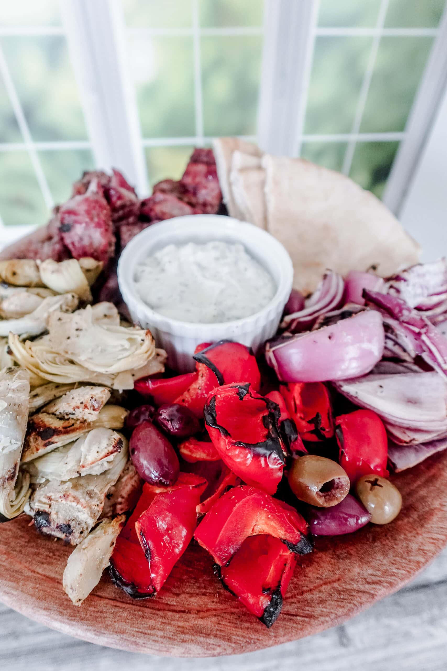 A wooden platter with grilled red peppers, artichokes, olives, red onions, pita bread, and grilled meat, surrounding a small bowl of creamy white dip, set in front of a bright window.
