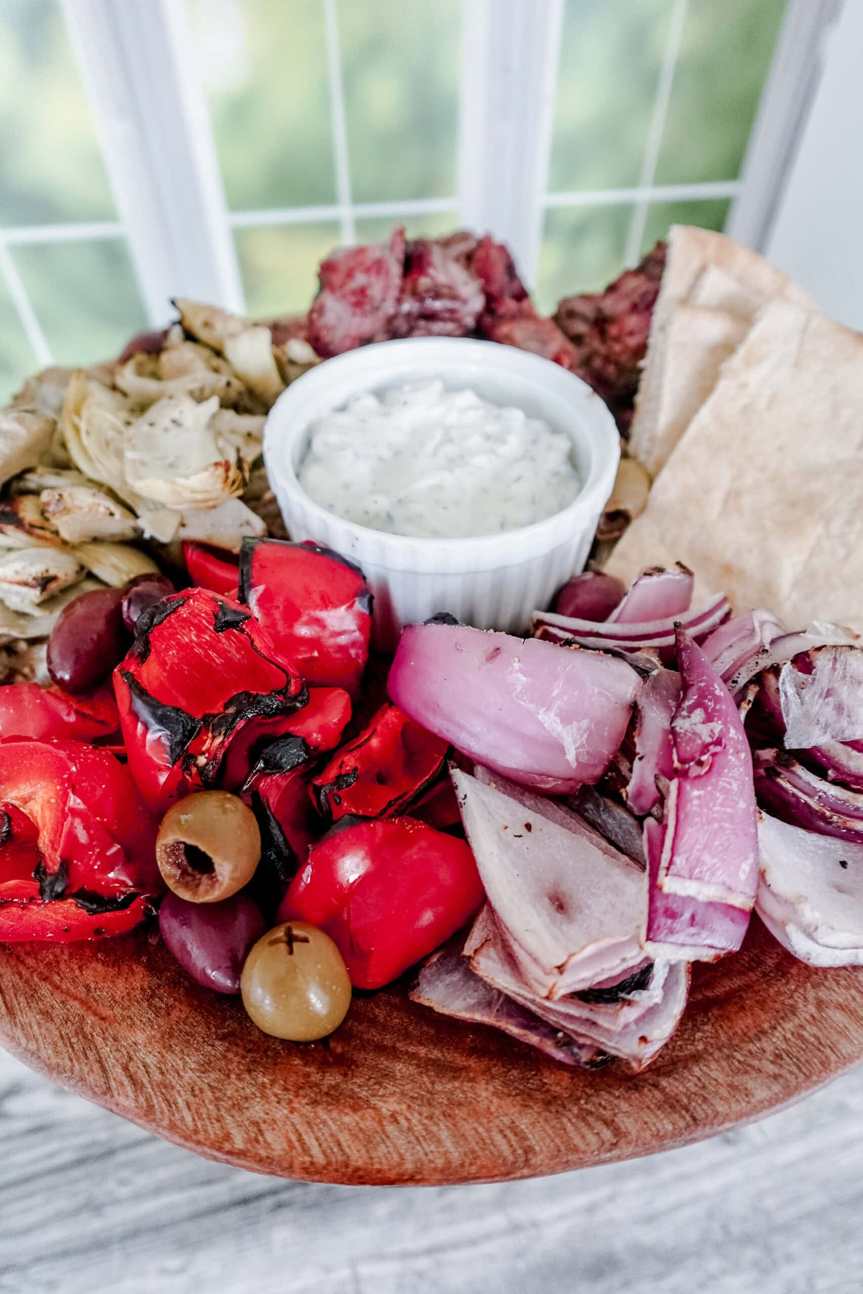 A wooden platter with grilled red peppers, red onions, olives, artichokes, pita bread, and a small bowl of creamy white dip in the center, set near a window with a blurred green outdoor view.
