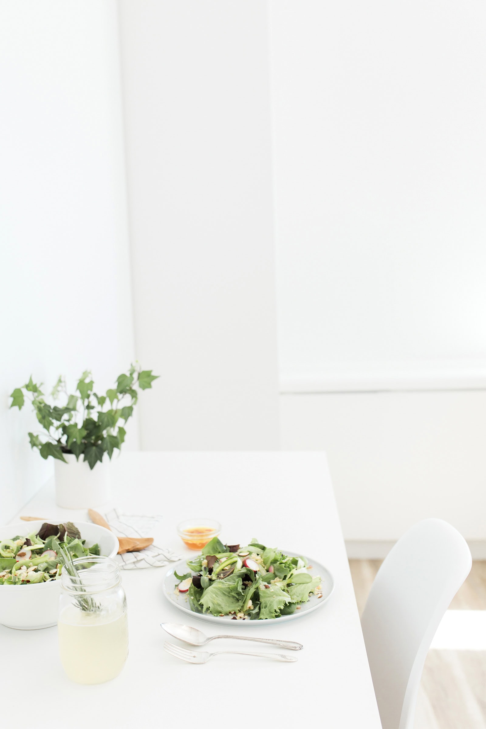 A bright, minimalistic dining table with a bowl and plate of fresh green salad, a glass of lemonade with herbs, utensils, a small dish, and a leafy green plant in the background.
