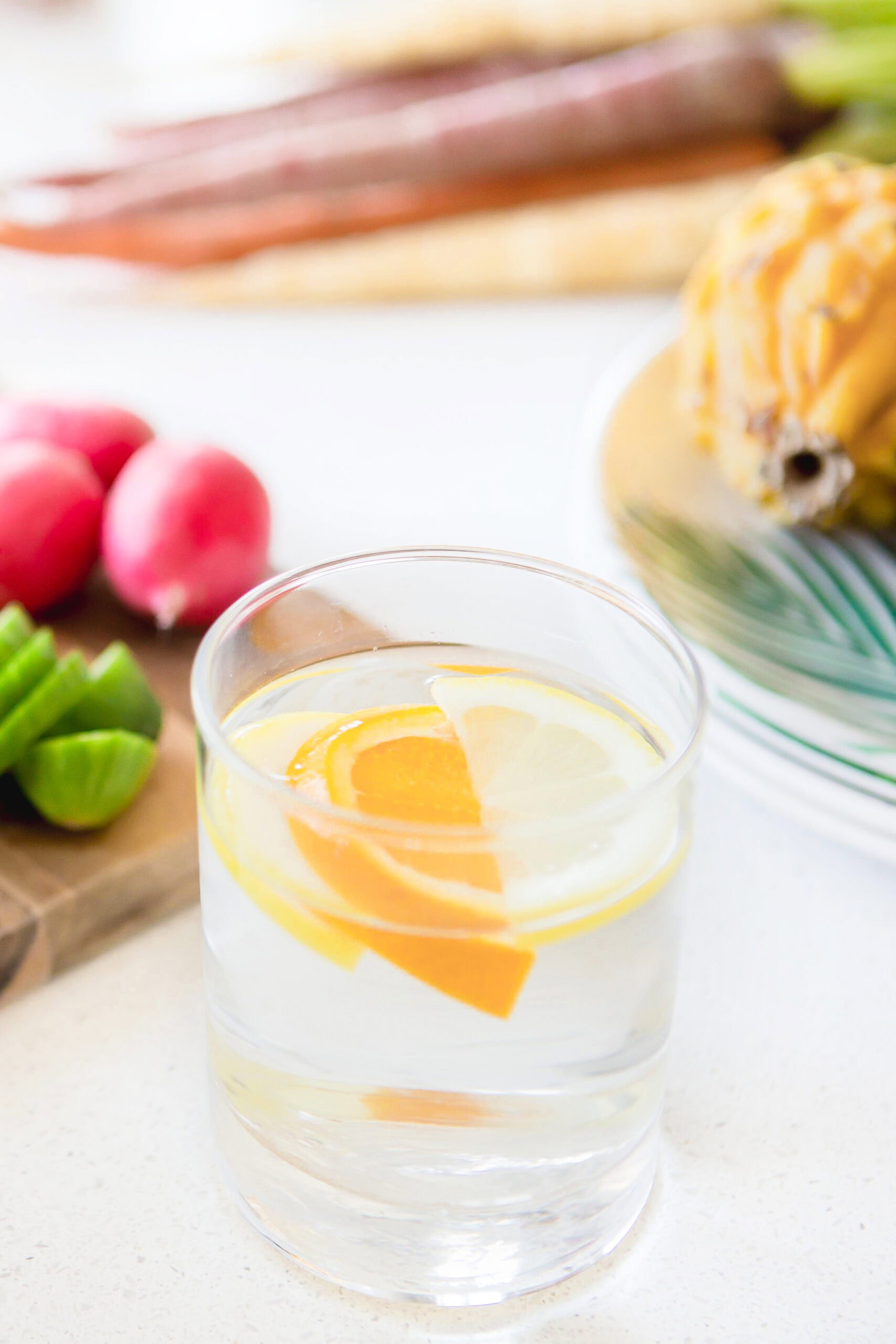 A clear glass of water with lemon and orange slices sits on a white surface, surrounded by fresh fruits and vegetables, including radishes, cucumbers, and carrots.