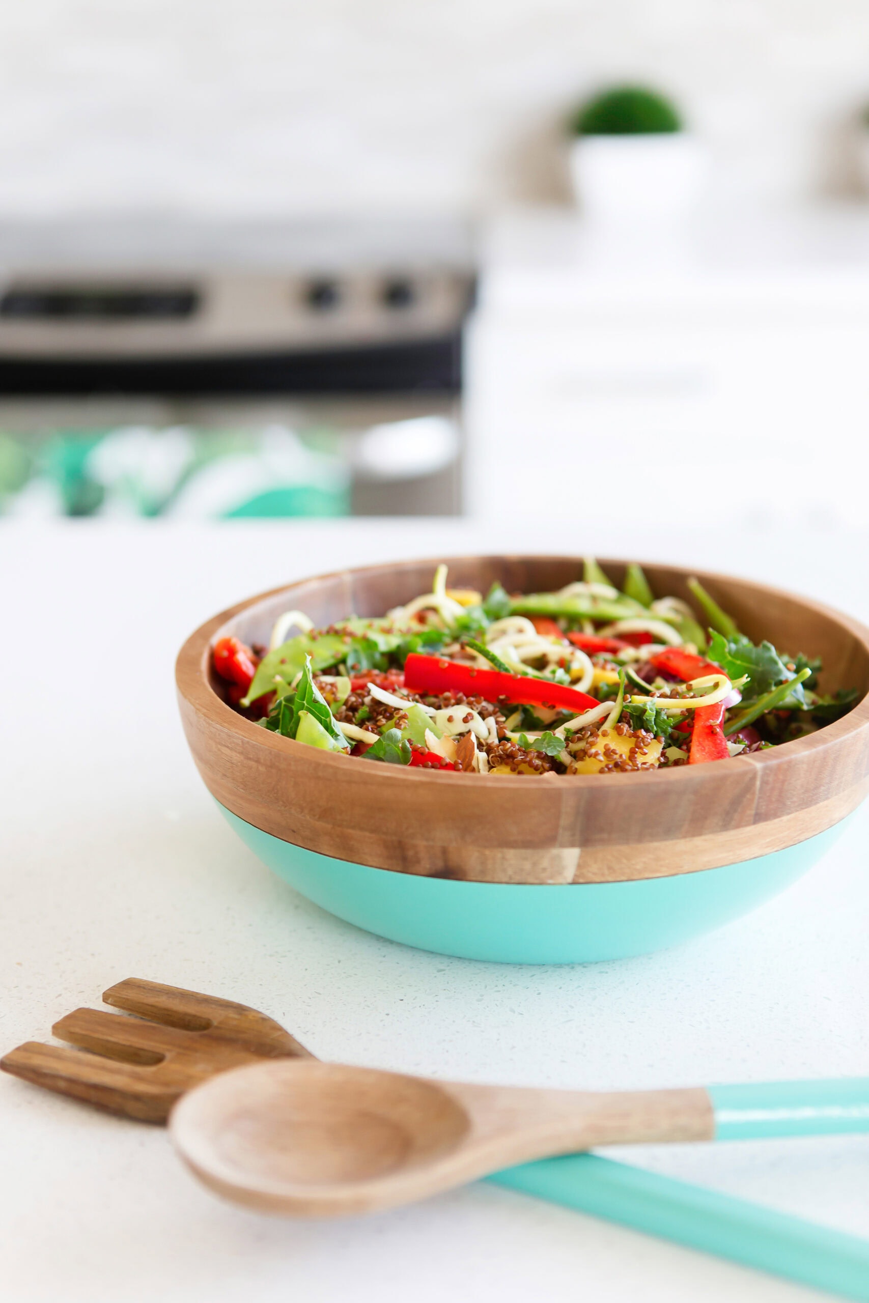 A vibrant salad with greens, sliced red peppers, and sprouts is served in a wooden bowl with a turquoise base, sitting on a white countertop next to wooden salad servers. A kitchen stove is blurred in the background.