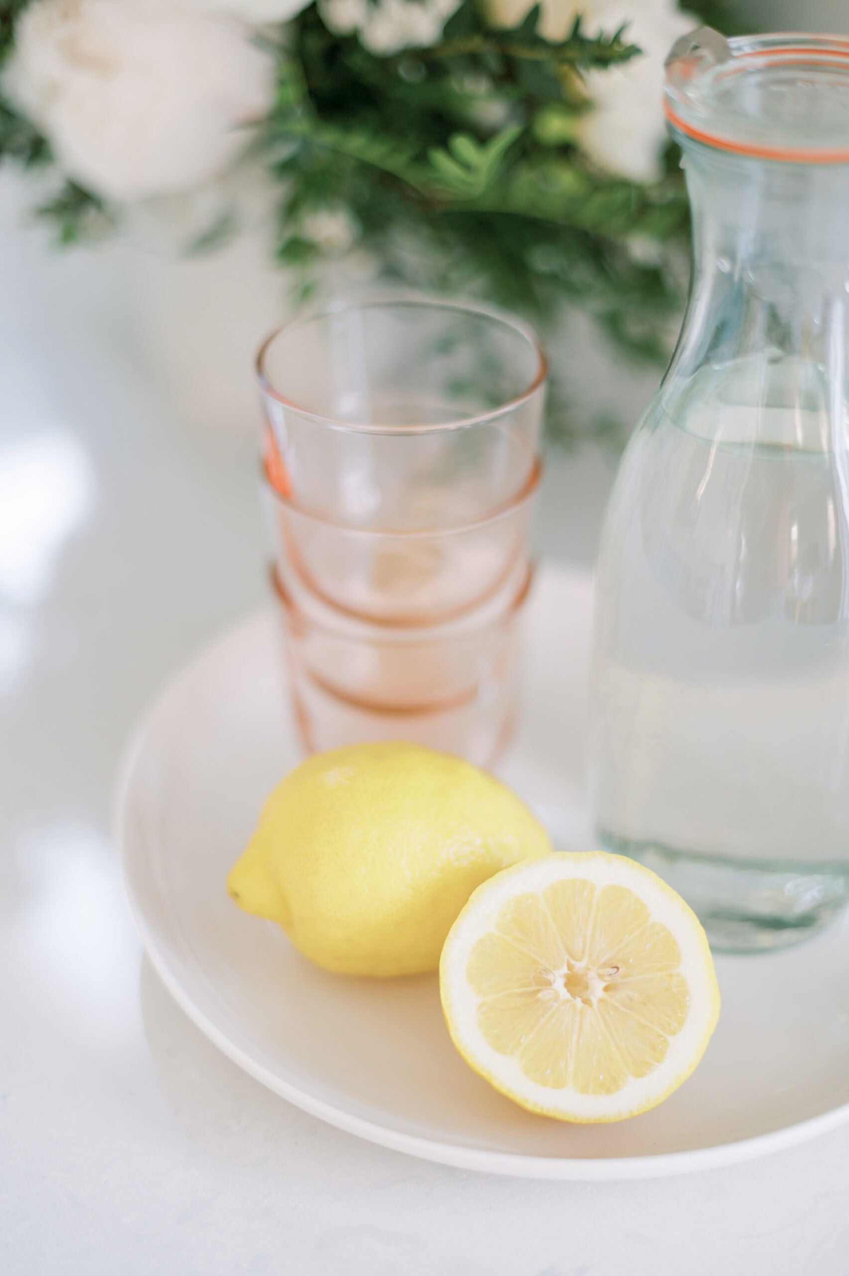 A lemon and a halved lemon sit on a white plate beside a glass carafe of water and stacked glasses, with blurred green leaves and white flowers in the background.