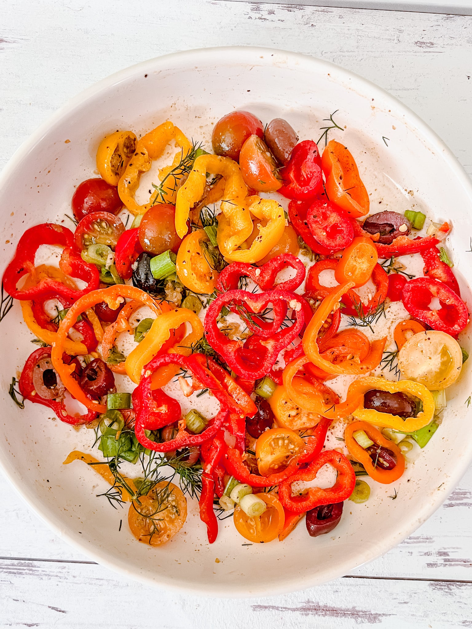 A white bowl filled with a colorful salad of sliced red, yellow, and orange bell peppers, cherry tomatoes, green onions, and fresh dill brings a taste of the Snug and Cozy Life to your table on a light wooden surface.