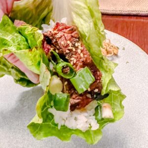 A hand holding a lettuce wrap filled with white rice, marinated beef, chopped green onions, and sesame seeds on a plate.