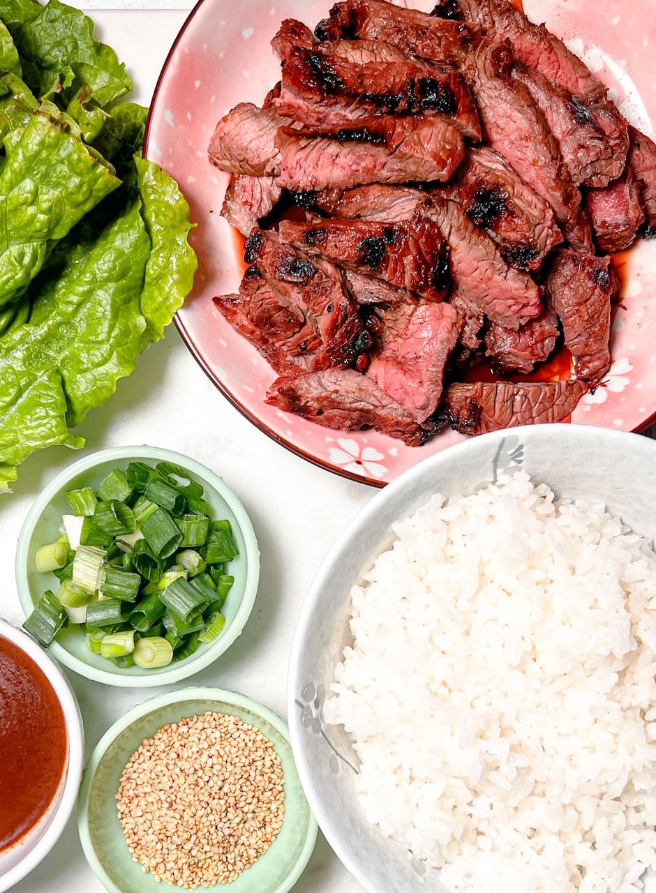 An overhead shot of a bowl of sliced grilled beef, a bowl of white rice, fresh lettuce leaves, chopped green onions, sesame seeds, and a small bowl of sauce arranged on a white surface.