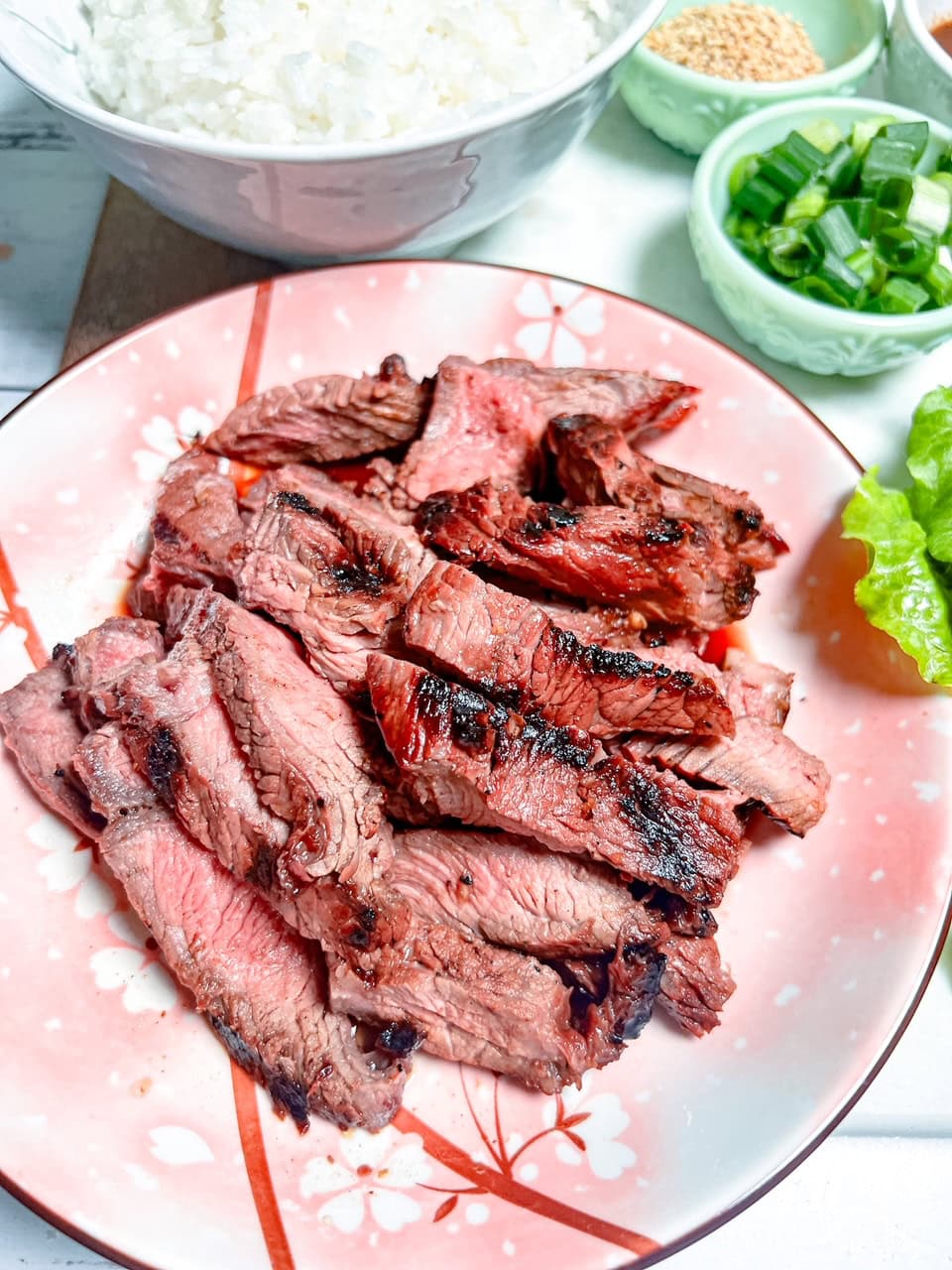 Sliced grilled steak served on a pink plate with a floral design, with bowls of rice, sesame seeds, chopped green onions, and lettuce in the background.