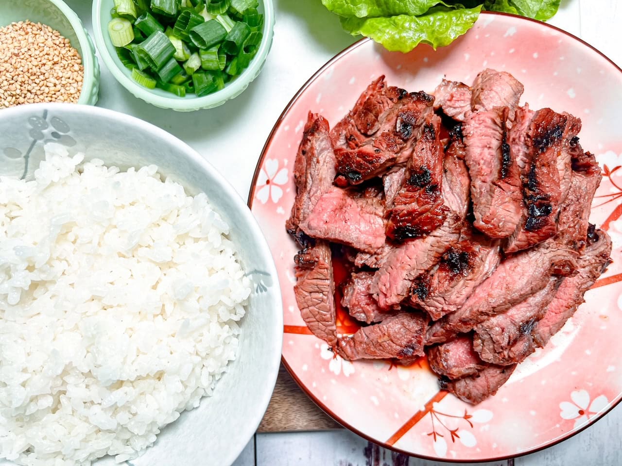 An overhead shot of a plate of grilled sliced beef on a pink cherry blossom-patterned dish, served with a bowl of white rice, green onions, sesame seeds, and fresh lettuce leaves on the side.