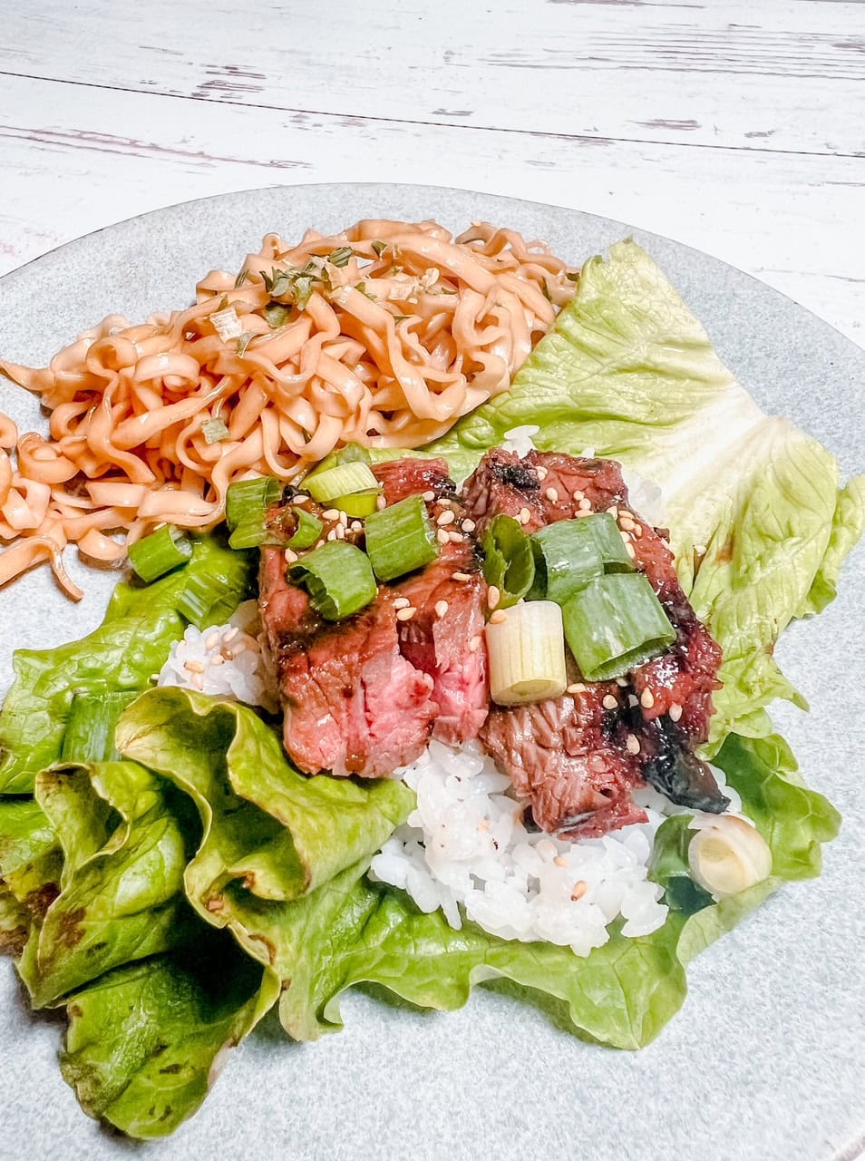 A close up of a plate with sliced steak topped with green onions and sesame seeds on a bed of rice and lettuce, served alongside a portion of saucy noodles.