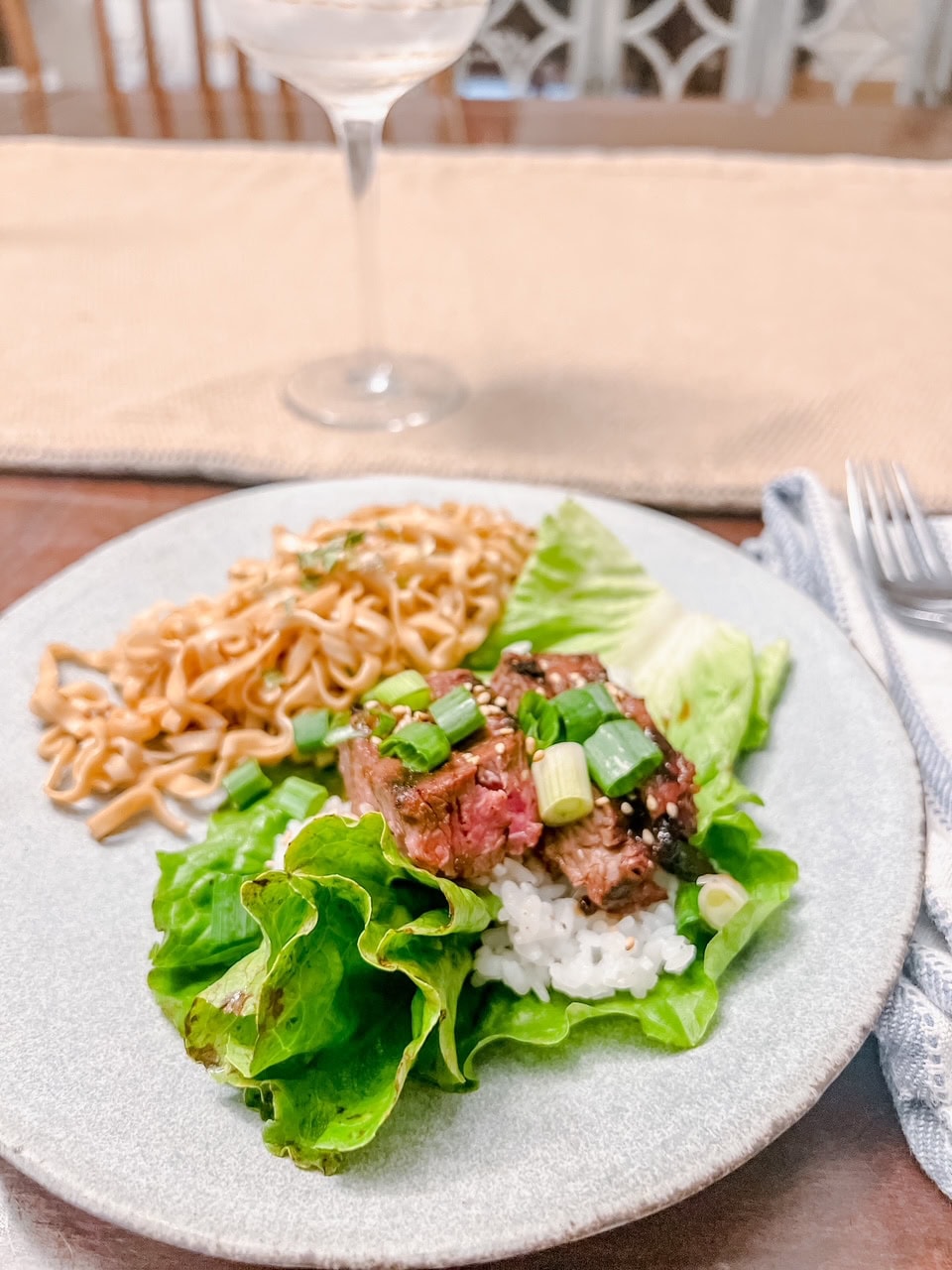 A plate of food with lettuce wraps filled with rice, sliced steak, and green onions, served alongside a portion of noodles. A glass of water and utensils are visible in the background.