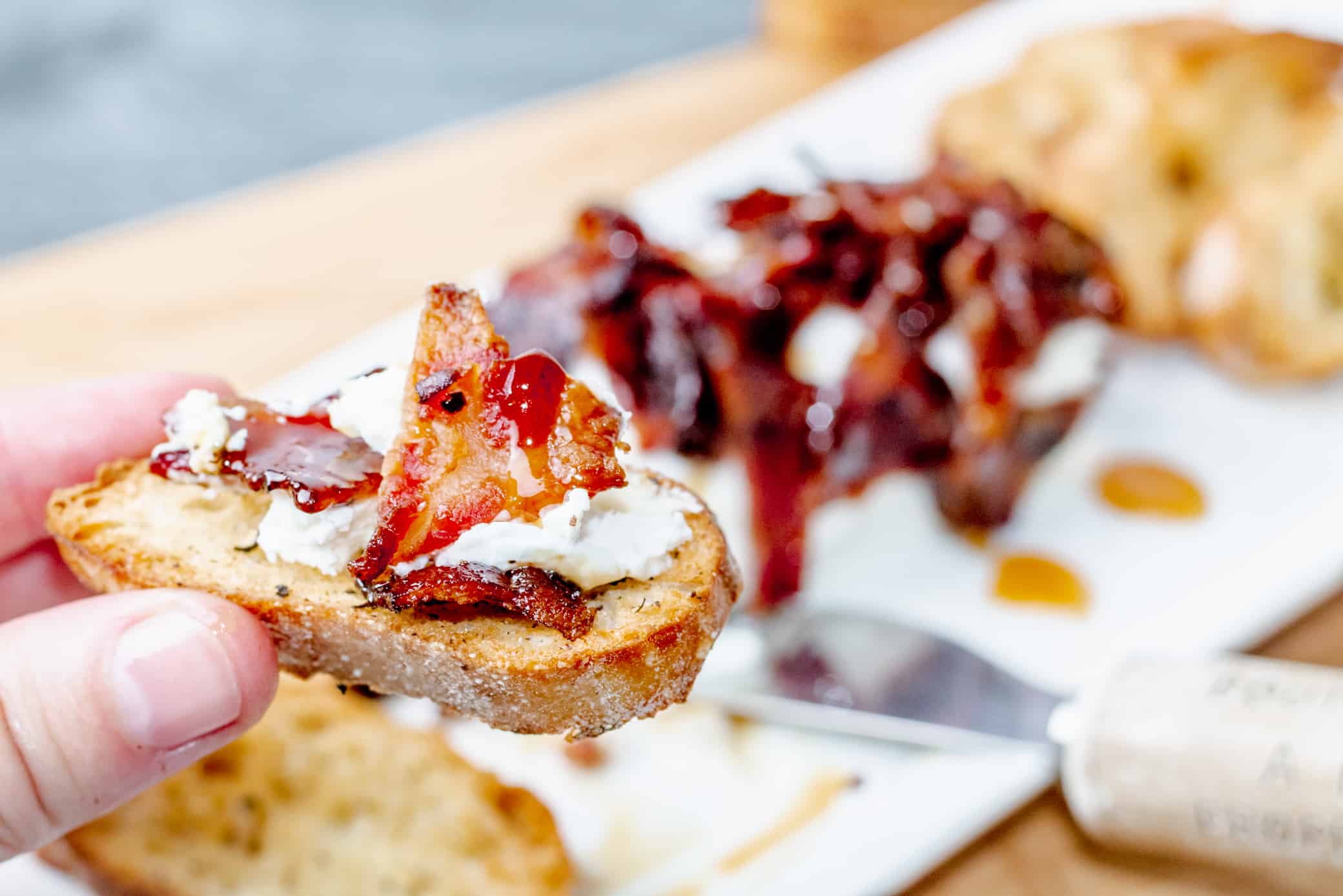 A hand holds a slice of toasted bread topped with creamy cheese and caramelized bacon, perfect for a Snug and Cozy Life. More topped bread slices and a cheese knife are blurred in the background on a white plate.