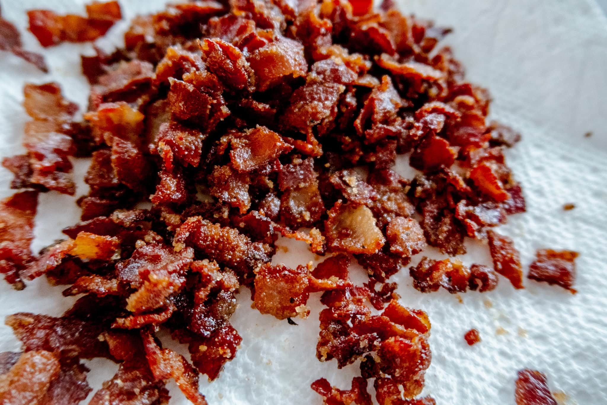 Close-up of crispy, crumbled bacon pieces resting on a white paper towel, showing their golden brown color and crunchy texture.