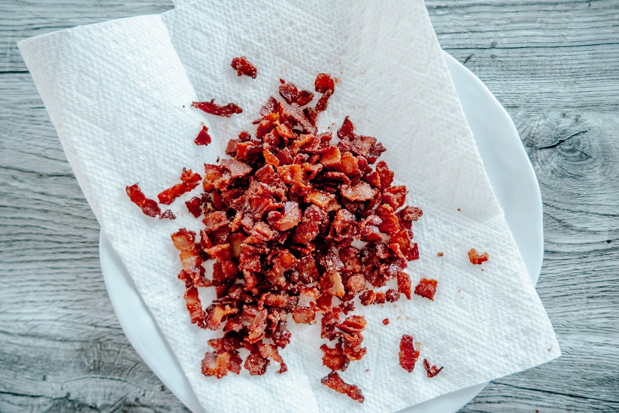 Chopped, cooked bacon pieces resting on a white paper towel atop a white plate, placed on a gray wooden surface.