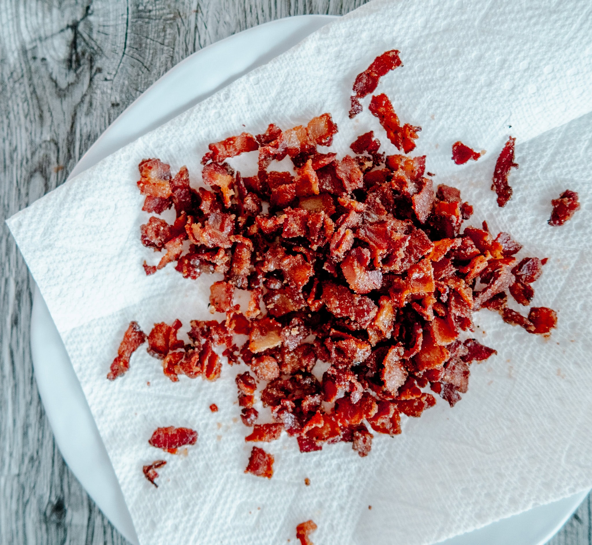 Chopped cooked bacon pieces are spread out on a paper towel, which is placed on a white plate atop a rustic wooden surface.