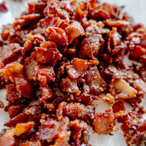 Close-up of crispy, chopped bacon pieces resting on a white paper towel, showing a mix of crunchy textures and rich, reddish-brown color.