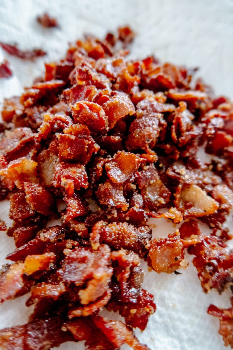 Close-up of crispy, chopped bacon pieces resting on a white paper towel, showing a mix of crunchy textures and rich, reddish-brown color.