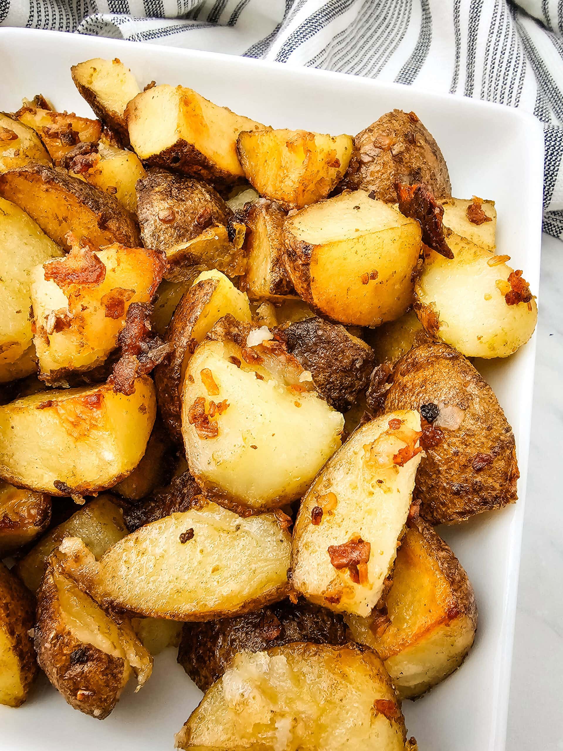 A close-up of crispy roasted potato wedges with bits of seasoning and bacon on a white rectangular plate, with a striped towel in the background.