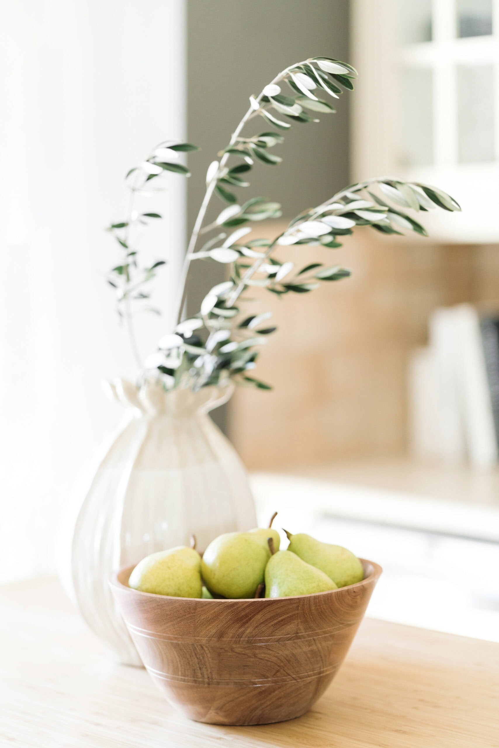 A wooden bowl filled with green pears sits on a light kitchen countertop. Behind it, a white ceramic vase holds leafy green branches, with soft natural light brightening the scene.