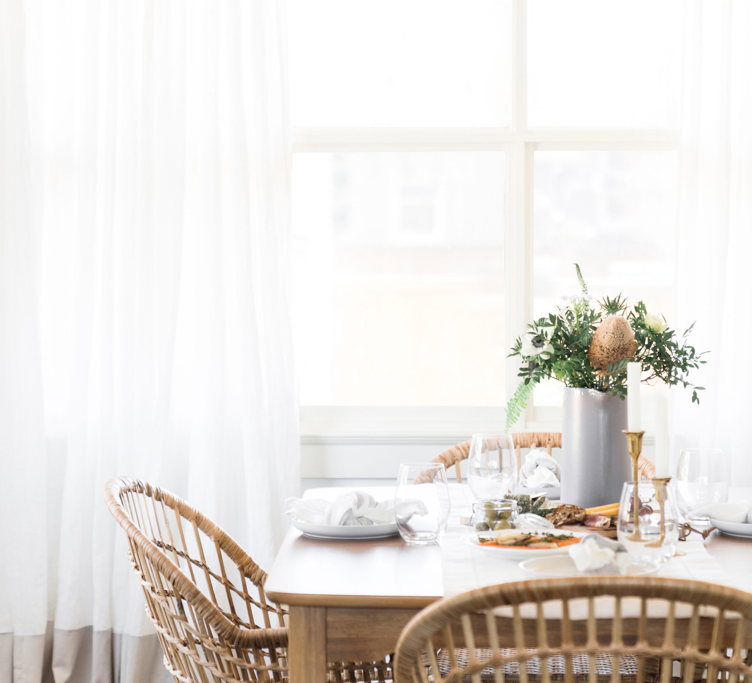 A bright dining area with wicker chairs around a wooden table set with plates, glasses, napkins, and a tall vase of green and brown flowers. Sunlight streams through large white-curtained windows in the background.