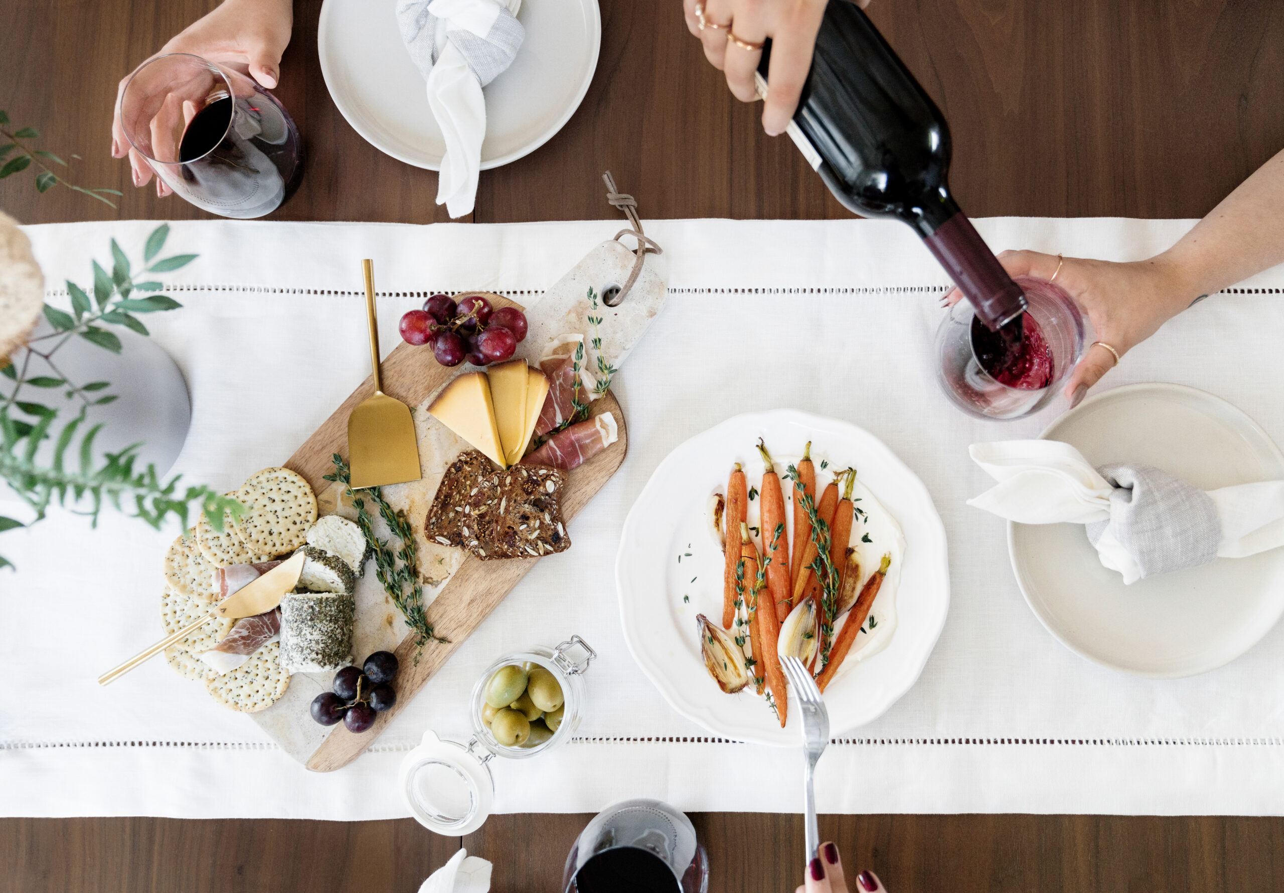 Overhead view of a dining table with a cheese and fruit board, roasted carrots on a plate, olives, and people pouring red wine and holding glasses, set on a white table runner.