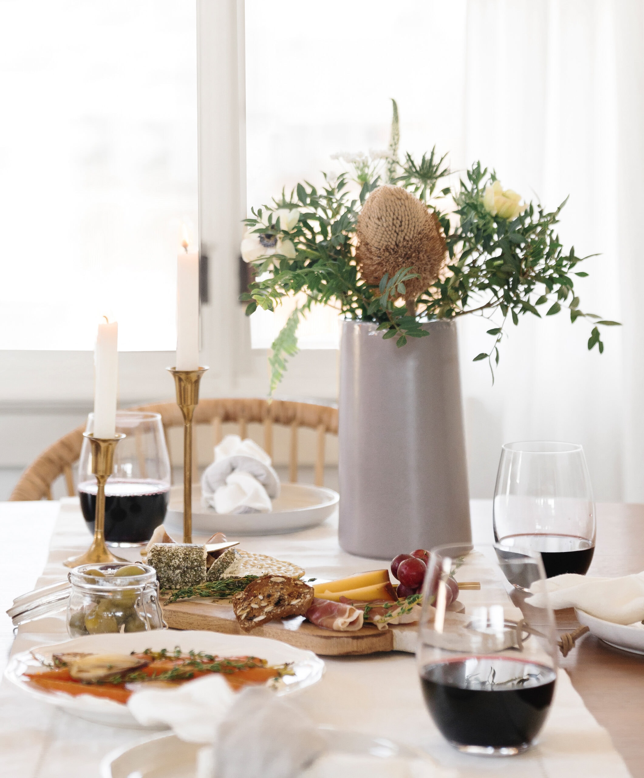 A dining table set with plates of appetizers, glasses of red wine, lit candles in holders, and a tall vase with assorted greenery and flowers, in a bright, airy room.