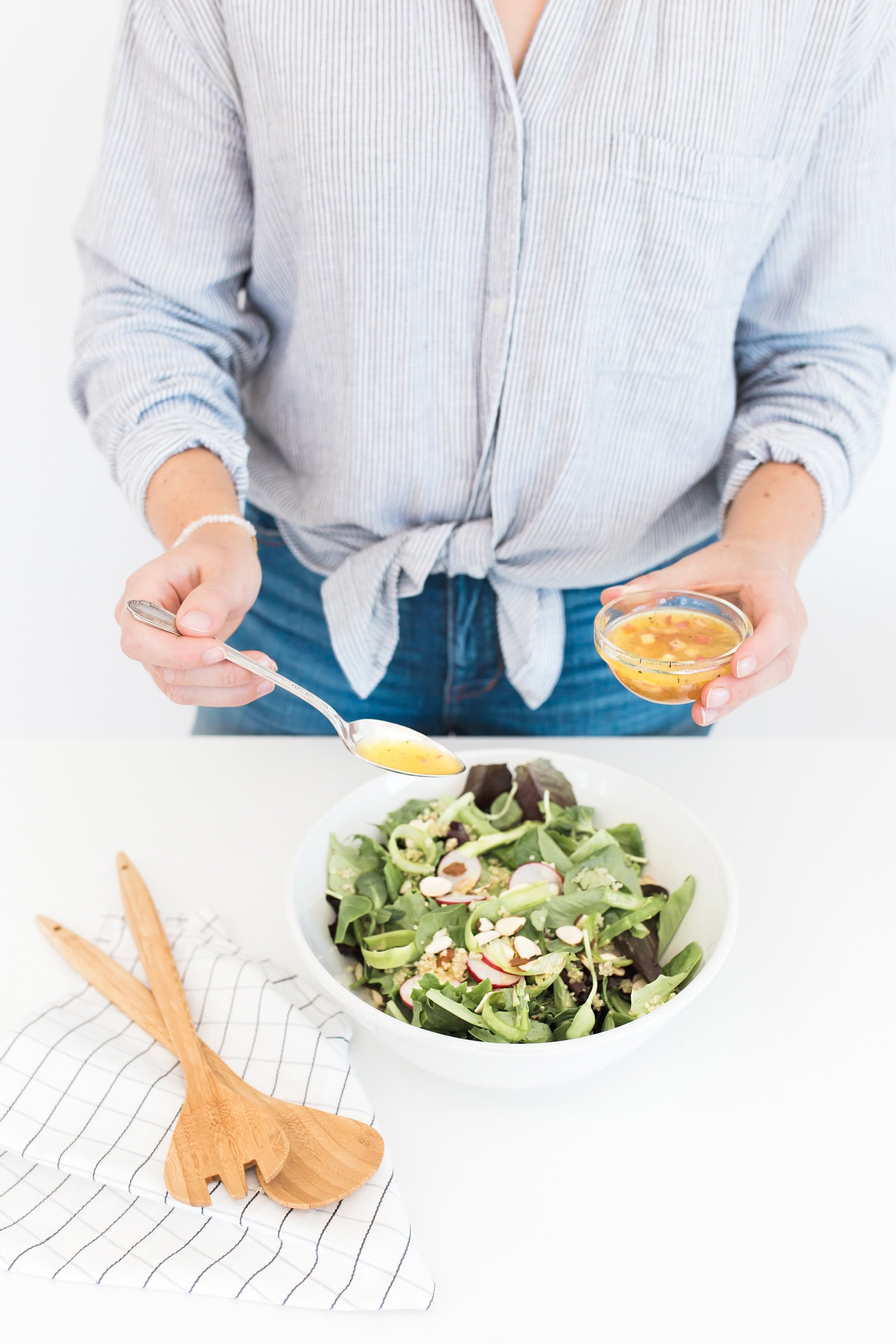 A person in a striped shirt is drizzling dressing onto a bowl of fresh salad with greens, radishes, and avocado. Wooden salad servers rest on a cloth beside the bowl on a white table.