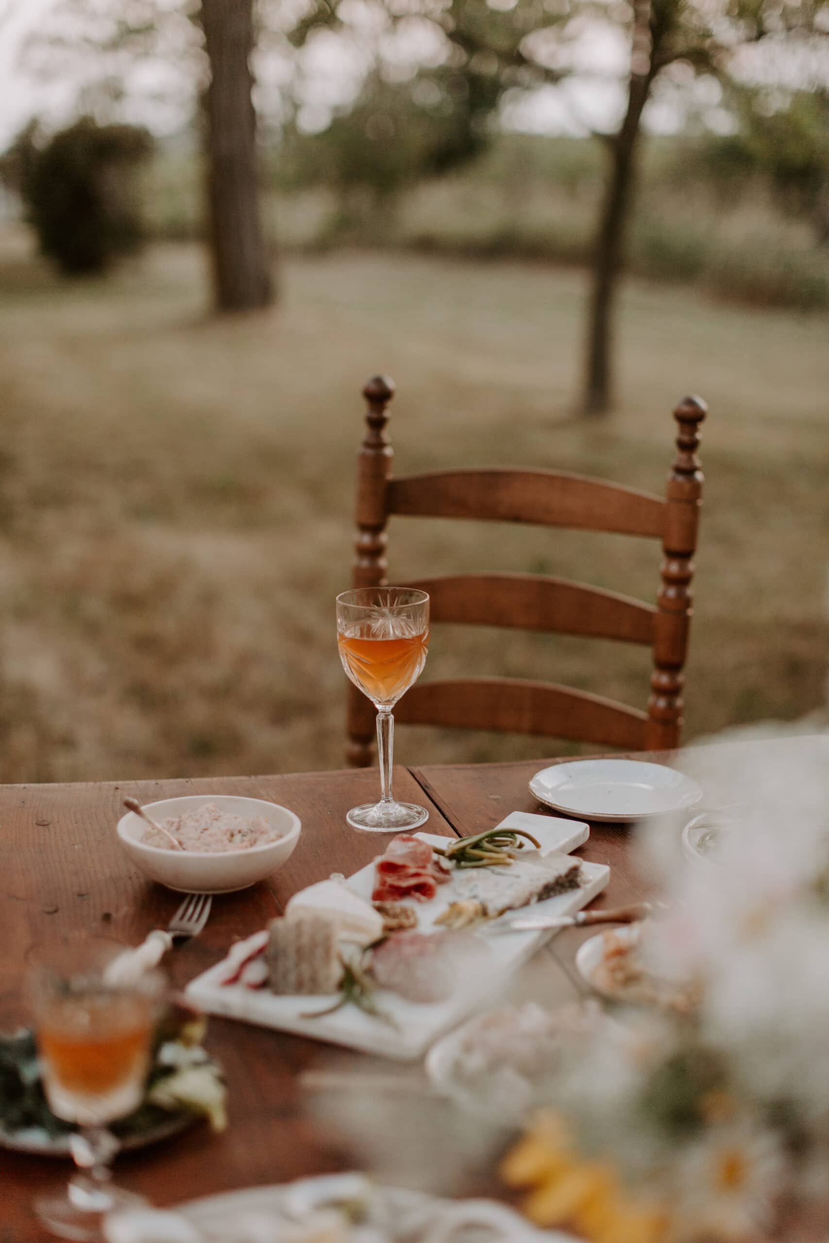 A wooden chair sits at an outdoor table set with a glass of orange-colored drink, a bowl, and plates of assorted appetizers, with trees and grass visible in the blurred background.