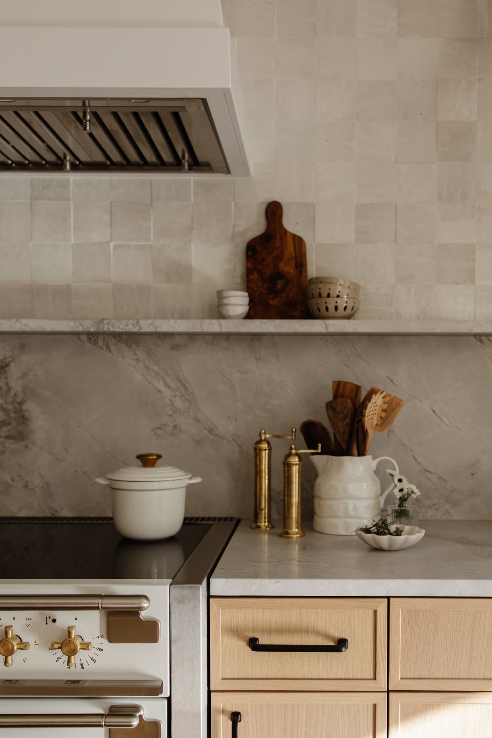 A modern kitchen with light wood cabinets, a marble backsplash, brass hardware, and utensils in a white pitcher. A white pot sits on the stove, and decorative items are arranged on a shelf above the counter.